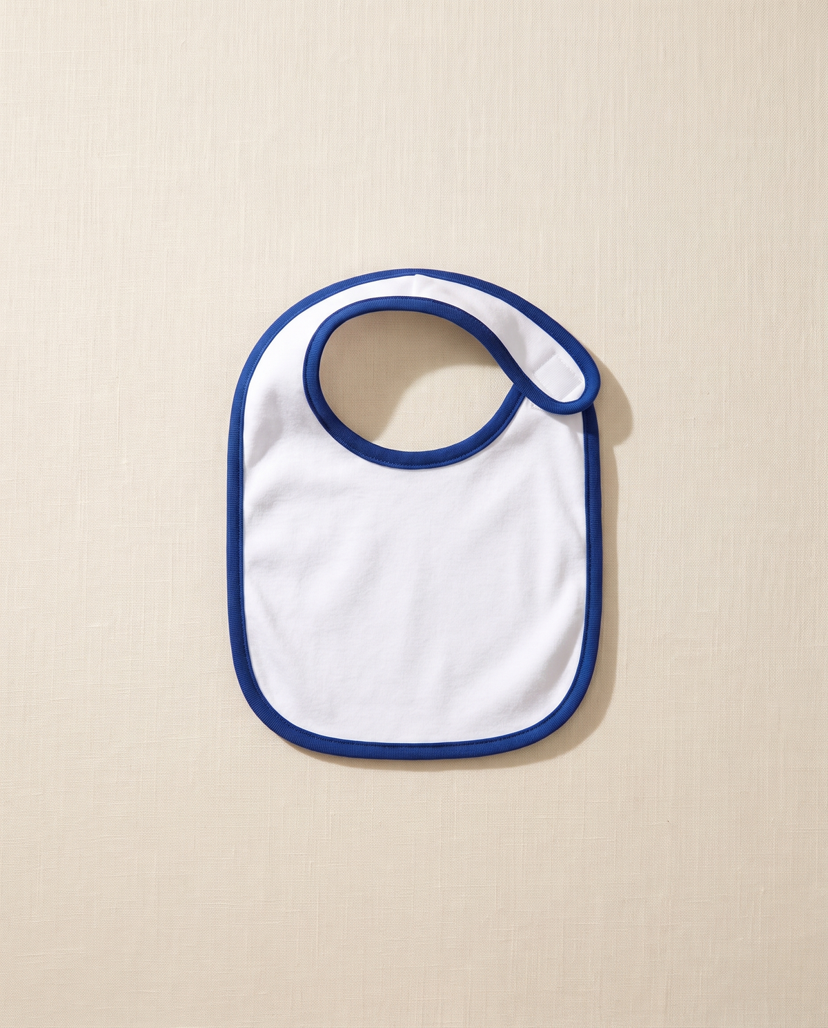 A blank white infant jersey bib with royal blue binding, laid flat on warm off-white linen