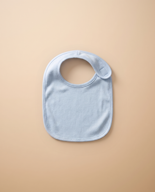 A blank light blue infant jersey bib, laid flat on warm off-white linen