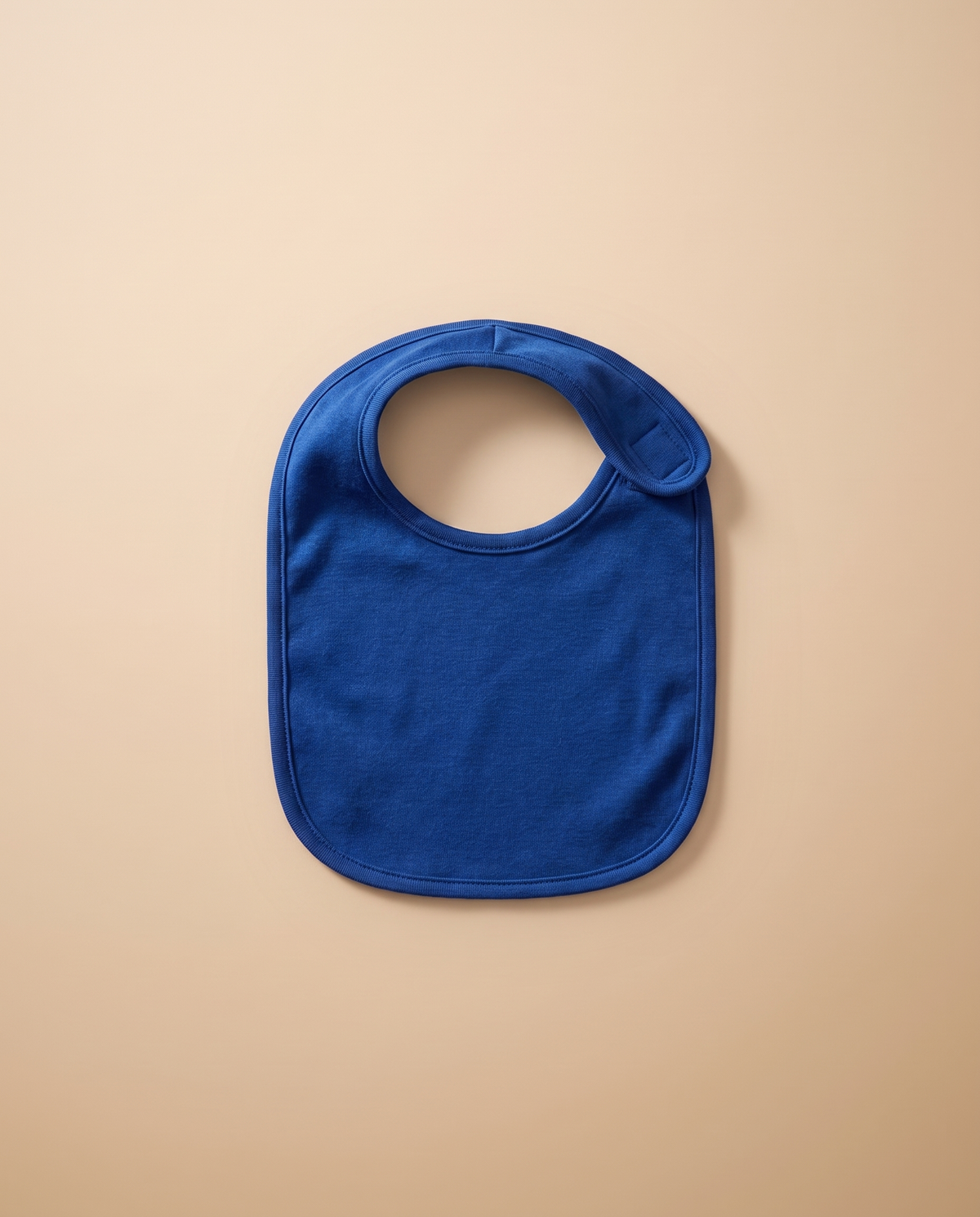 A blank royal blue infant jersey bib, laid flat on warm off-white linen