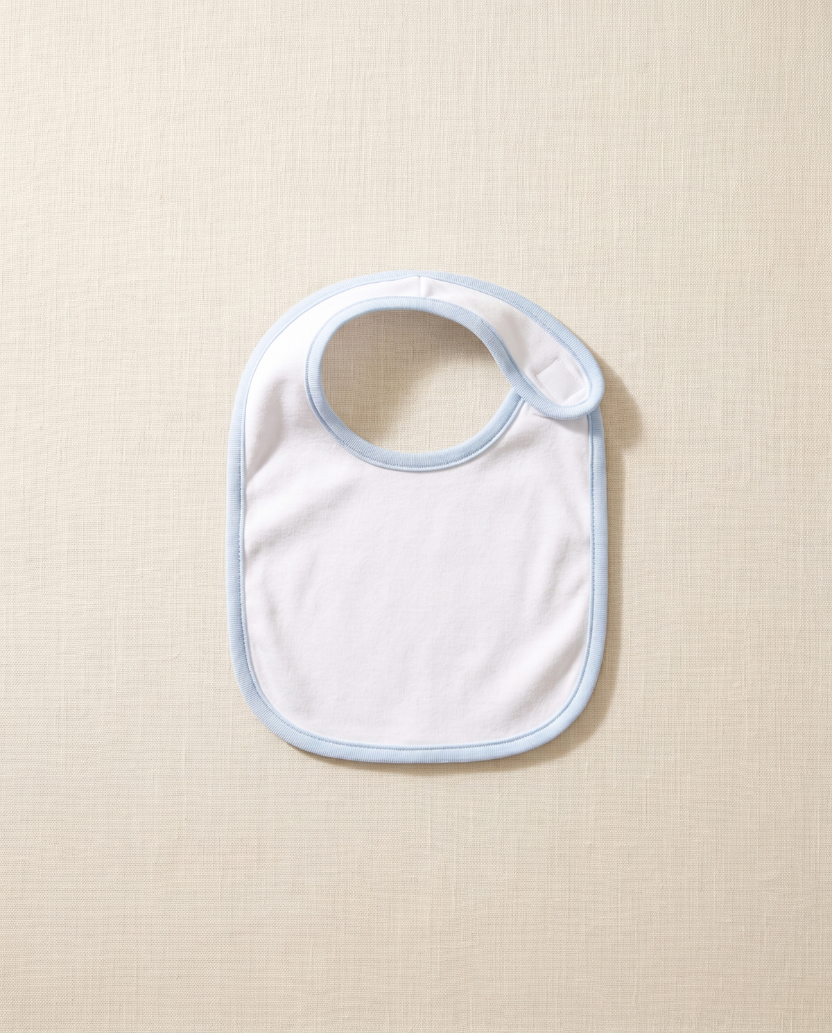 A blank white infant jersey bib with light blue binding, laid flat on warm off-white linen