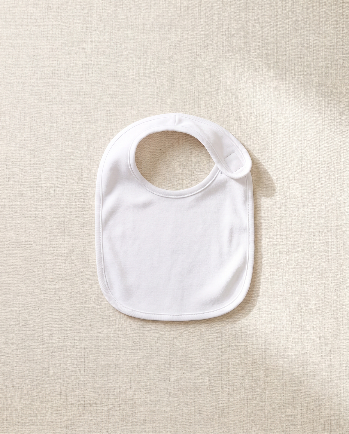 A blank white infant jersey bib, laid flat on warm off-white linen